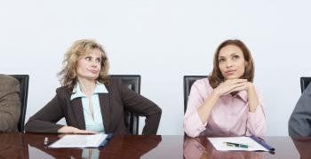Two women sitting in a conference room, one rolling her eyes at the other.